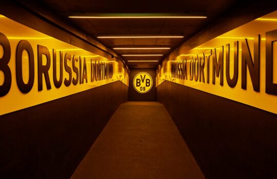 Long tunnel with yellow-lit walls bearing BORUSSIA DORTMUND, leading to a round BVB emblem at the far end.
