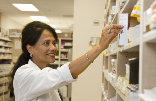 A pharmacist in a white coat reaching for a medication box on a shelf