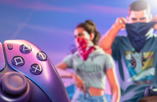 Close-up of a purple game controller in the foreground; two masked people in a neon-lit background.