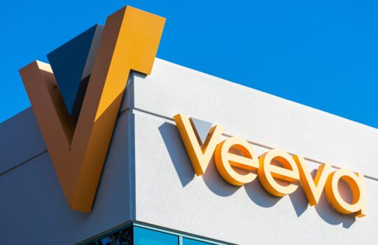 Veeva logo in orange letters on a white wall against a clear blue sky.