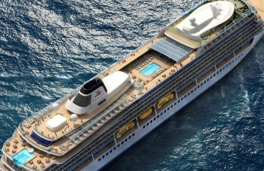 Aerial view of a large cruise ship at sea, with multiple sunlit decks, a central pool, and yellow lifeboats along the side.
