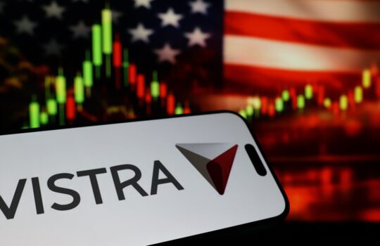 Vistra logo on a white sign with a red and gray triangular symbol, held in front of a blurred stock chart and American flag background.