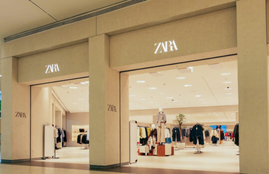 ZARA storefront inside a shopping mall with clothing on racks visible through glass walls and beige pillars around.