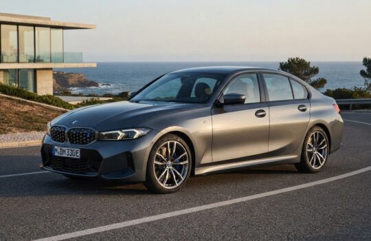 Gray BMW 3 Series sedan parked on a coastal road with the sea and a modern glass house in the background.