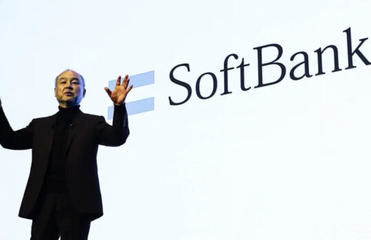 Male presenter on stage gesturing with both hands in front of a large SoftBank logo behind him on a white backdrop.