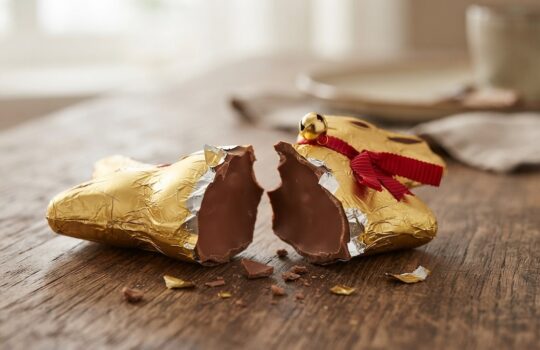 Broken milk chocolate candy in gold foil with a red ribbon, split in two on a wooden table.