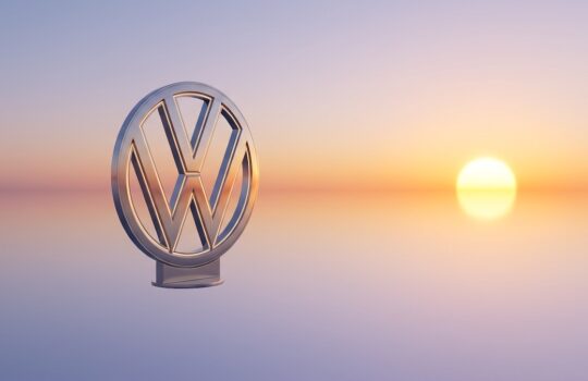 Metal Volkswagen logo emblem mounted on a stand, with a serene sunset over calm water in the background