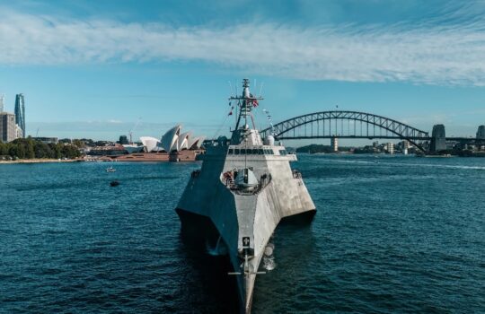Front view of a gray warship in Sydney Harbour with the Opera House and Harbour Bridge in the distance on a sunny day.