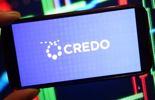 Smartphone held horizontally showing a blue screen with the white 'CREDO' logo and dotted circular mark against a colorful neon background.