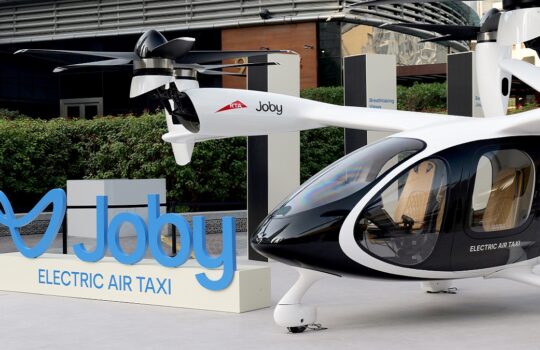 White electric air taxi with two rotor props parked beside a blue 'Joby Electric Air Taxi' sign in an urban plaza.