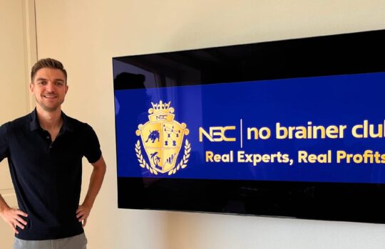 Smiling man in a navy polo stands next to a wall-mounted screen displaying the blue 'no brainer club' logo and slogan: Real Experts, Real Profits.