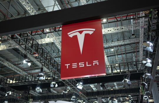 Red Tesla banner with white T logo hanging from ceiling trusses at an indoor exhibition hall