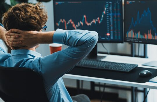 Person in a blue shirt sits back in a chair with hands behind their head, facing two monitors showing stock market charts a trading desk scene.