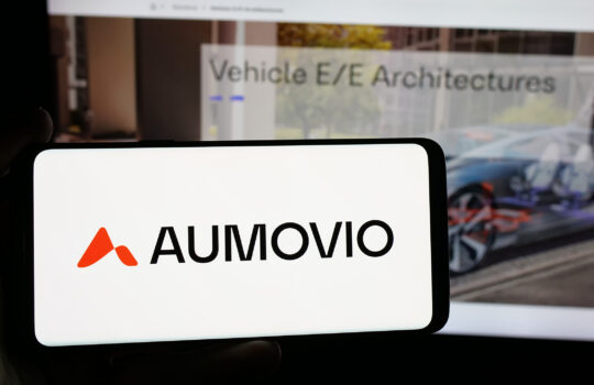 Close-up of a white card displaying the AUMOVIO logo in orange with bold black text; a blurred computer screen is in the background showing the same branding.