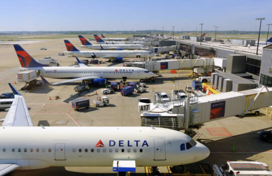 Several Delta aircraft parked at gates with jet bridges and ground crew equipment on the tarmac at an airport. 2. Aerial view of Delta planes lined up at terminal gates, boarding bridges connected. 3. Delta Air Lines airplanes at an airport gate area, with service vehicles nearby. 4. Row of white Delta jets at an airport terminal, awaiting boarding at the jet bridges. 5. Delta jet at the gate with walkway and ground vehicles around.