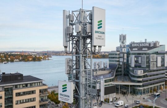 Ericsson telecommunications tower with multiple antenna panels in the foreground, overlooking a river and modern campus buildings.