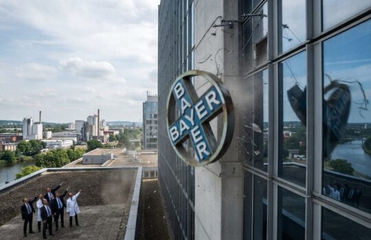Group of professionals on a rooftop look up at a Bayer circular logo mounted on a glass-building wall, with city and river in the background.