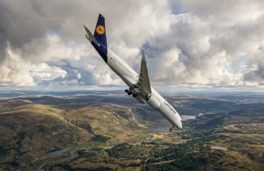 Lufthansa airliner climbs upward with landing gear extended over a valley of rolling hills, lakes, and winding rivers below.