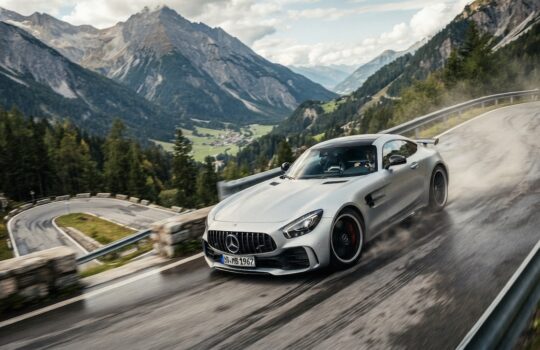 Silver sports car speeds along a winding mountain road with guardrails and forested slopes in the background.