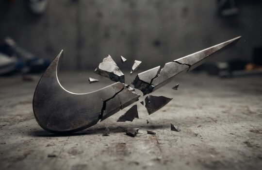 Broken metal Nike swoosh lying on a dusty concrete floor with shards scattered around it, as if shattered in a workshop moment.